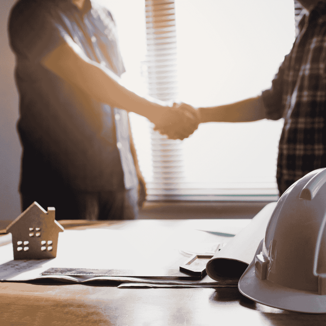 Zwei Personen geben sich die Hand während der Projektplanung, mit Bauhelm und Bauplänen auf dem Tisch – Umbau Expert